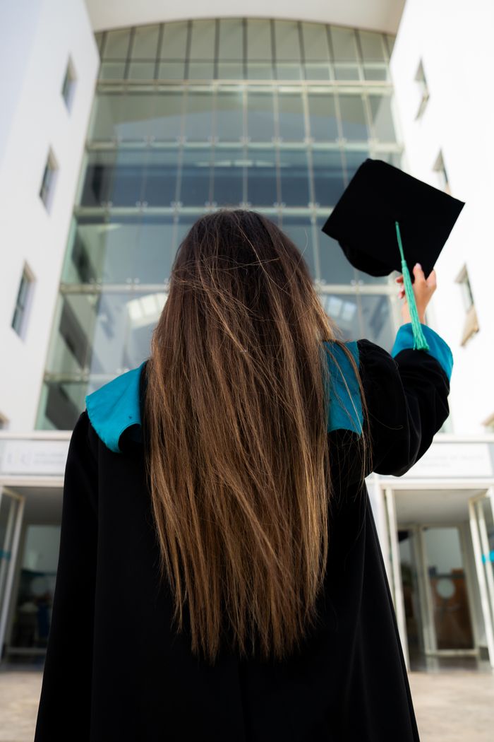 Young woman holding graduation cap outside modern university building, celebrating achievement.