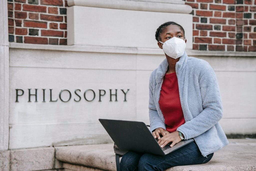 Anonymous African American female student of Philosophy faculty in casual clothes and face mask sitting near university building and working on laptop