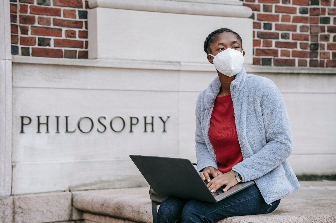 Anonymous African American female student of Philosophy faculty in casual clothes and face mask sitting near university building and working on laptop