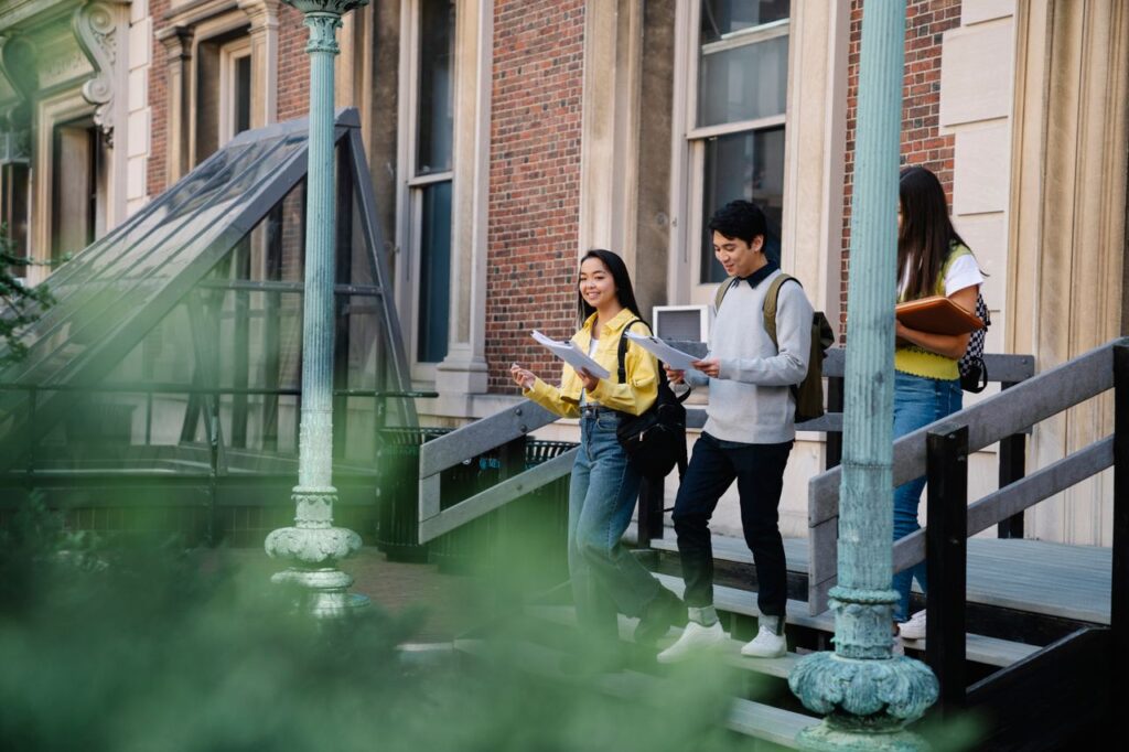 University students walking downstairs, holding papers, and enjoying a bright day.